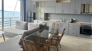 Una cocina moderna con armarios grises, una mesa de comedor de cristal, sillas de mimbre y vistas al mar a través de amplios ventanales.