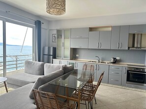 A modern kitchen with grey cabinets, a glass dining table, wicker chairs, and a view of the sea through large windows. - Emily - Bougainville Panorama (Sarandë)
