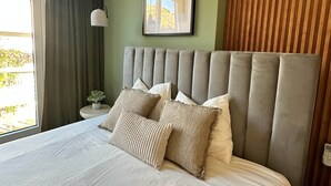 Een moderne slaapkamer met een groot bed, hoofdbord, een lijst met een kunstwerk, een raam met gordijnen en een nachtkastje met een lamp.