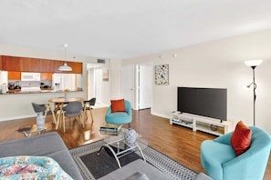 A modern living room with a sofa, armchair, and coffee table. There is a dining area with chairs and a table, and a kitchen with wooden cabinets and appliances. A flat-screen TV is mounted on the wall. - Brickell Bay Condo with Huge Balcony and Pool (Miami)