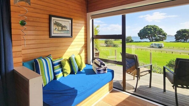 A room with a wooden wall, a blue sofa with striped cushions, a framed picture, and a view of a green landscape through a glass door. - Hahei Beachside Escape (Hahei)