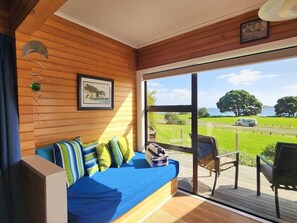 A room with a wooden wall, a blue sofa with striped cushions, a framed picture, and a view of a green landscape through a glass door. - Hahei Beachside Escape (Hahei)