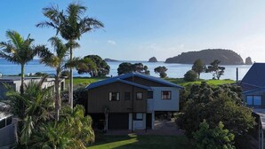 A modern house with a garage, surrounded by lush greenery and palm trees, overlooking a calm sea and distant islands. - Hahei Beachside Escape (Hahei)