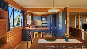 A compact kitchen with wooden walls, a central island, and a dining area with a table and chairs. - Hahei Beachside Escape (Hahei)