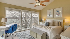Ein Schlafzimmer mit großem Fenster, einem Bett, einem Schreibtisch und einem Deckenventilator.