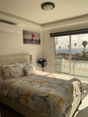 A bedroom with a bed featuring Paris-themed bedding, a ceiling fan, a painting of a couple with an umbrella, and a view of palm trees and the sea through large windows.