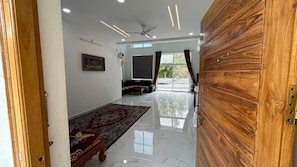 Un vaste espace de vie agrémenté d’une porte en bois, d’un ventilateur de plafond, d’une grande fenêtre avec des rideaux, d’un canapé et d’un tapis rouge à motifs.