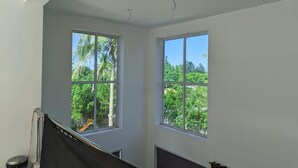 Una habitación con dos ventanas amplias, paredes blancas y vistas a espacios verdes.