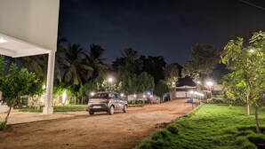Zona exterior bien iluminada con un camino de tierra, un SUV estacionado y una estructura de edificio blanco.