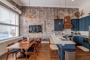 A modern kitchen with a brick wall, blue cabinets, a large island, and a dining area with wooden chairs and a table. - High-End Historic Loft | Premium DT Stay (Knoxville)