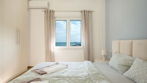 Quarto com cama, mesa de cabeceira com abajur, janela com cortinas e vista para o mar.