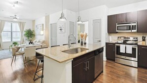 A modern kitchen with dark cabinets, a central island with a sink, a microwave, an oven, and a dining area with a table and chairs. - Zen 2B2B Next to Legacy West (Frisco)