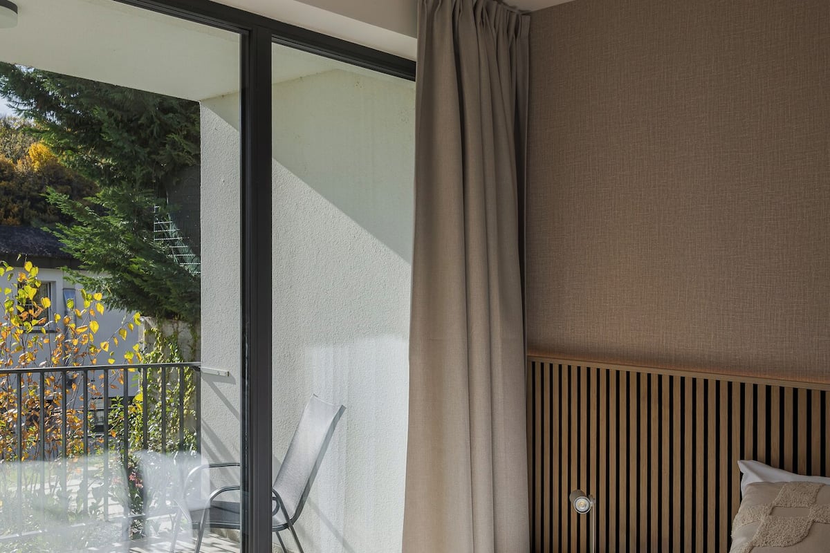 Une chambre d’hôtel dotée d’une grande porte-fenêtre coulissante donnant sur un balcon, d’un lit avec du linge de lit blanc, d’une table de chevet avec une lampe et d’une tête de lit en bois.