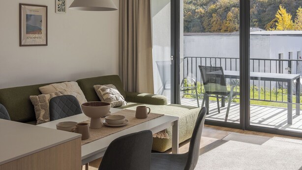 Un salon moderne avec un canapé, une table à manger et des chaises. Il y a une grande fenêtre donnant sur des arbres et un balcon.