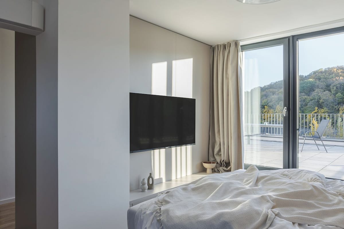 Une chambre moderne avec un grand lit, un téléviseur à écran plat, une plante en pot et une vue sur un balcon doté d’une balustrade.
