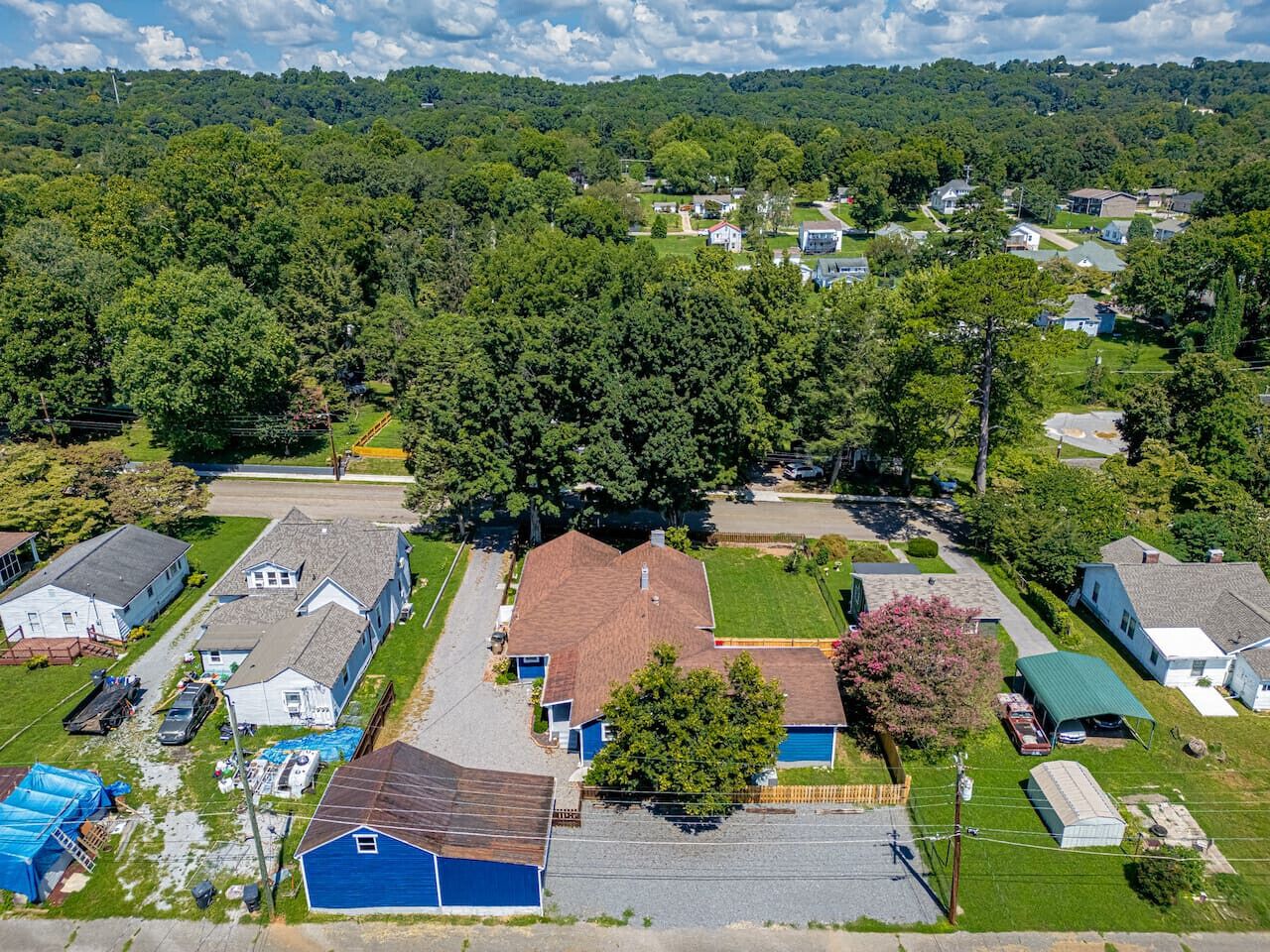 An aerial view of a residential area with houses, a large green field, and a dense forest in the background.