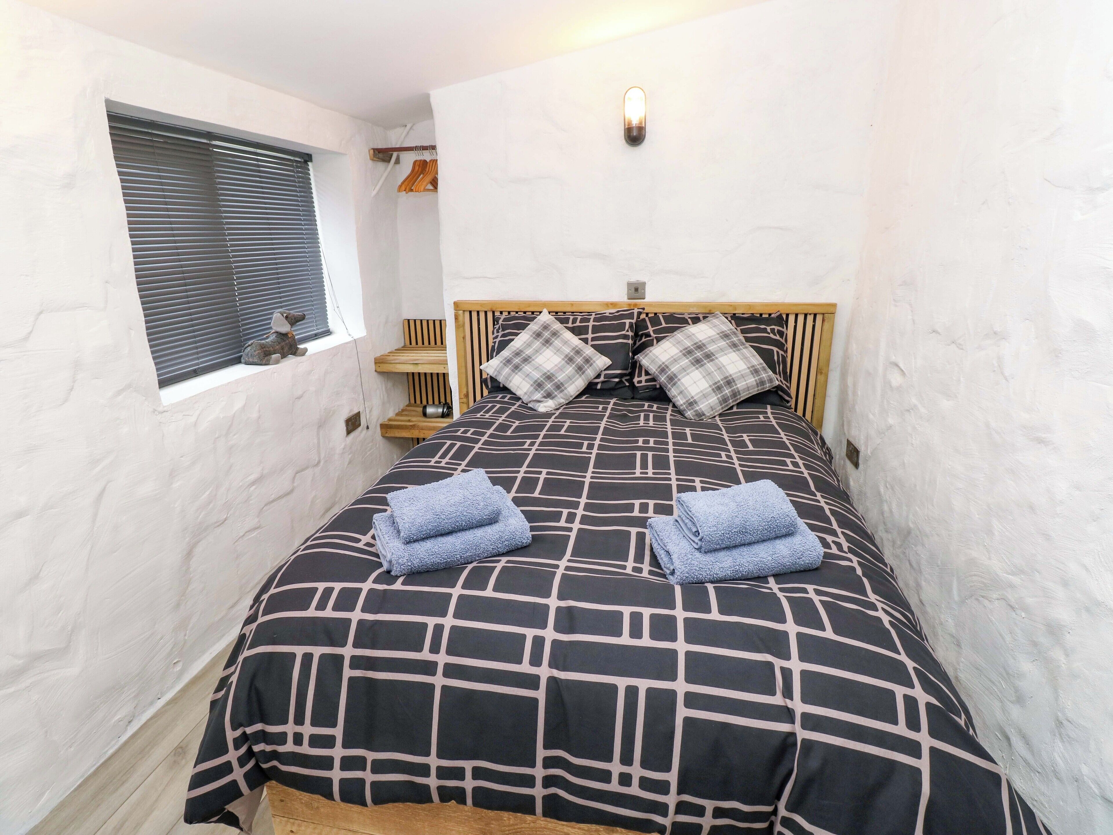 A narrow room with a bed, a window with blinds, a bedside table with a lamp, and a wall-mounted light fixture.