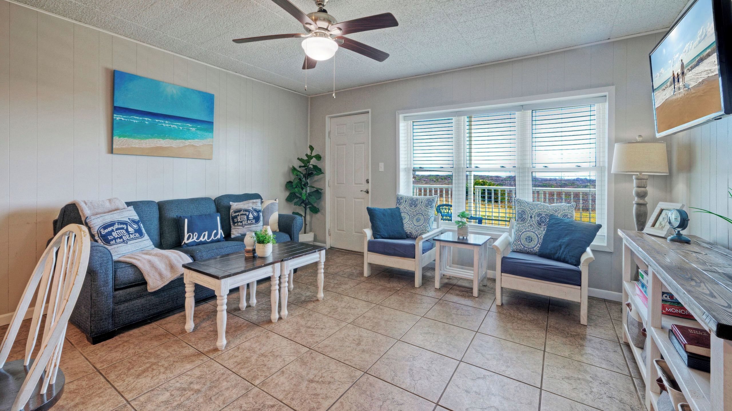 A living room with a ceiling fan, a large window with blinds, a sofa, a coffee table, and a bookshelf.