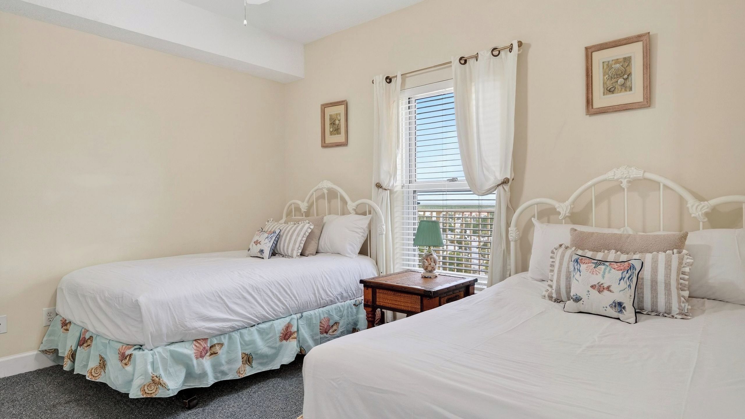 A bedroom with two beds, a window with curtains, a bedside table with a lamp, and framed pictures on the wall.