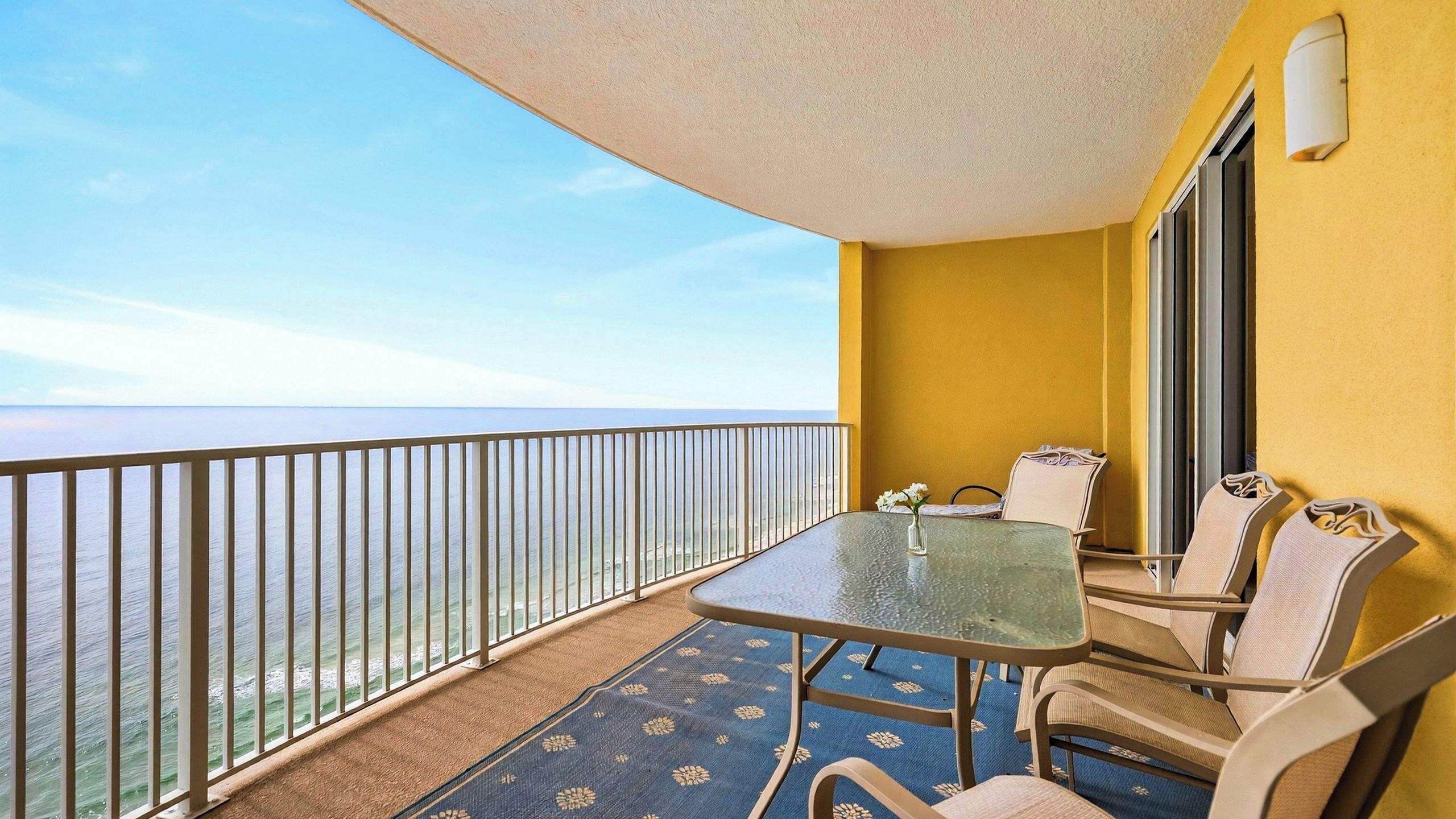 A balcony with a glass table and chairs, a view of the sea, and a yellow wall.