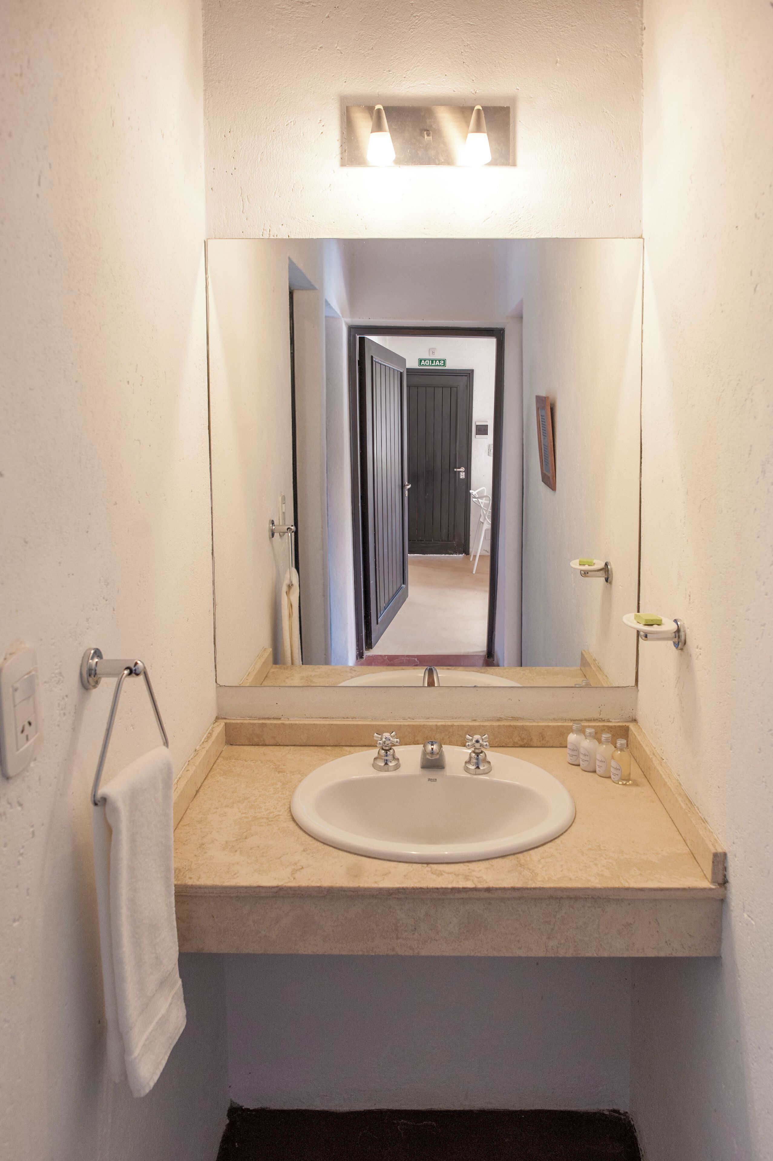 Un baño con un espejo grande, un lavamanos, un perchero para toallas y una lámpara sobre el espejo.