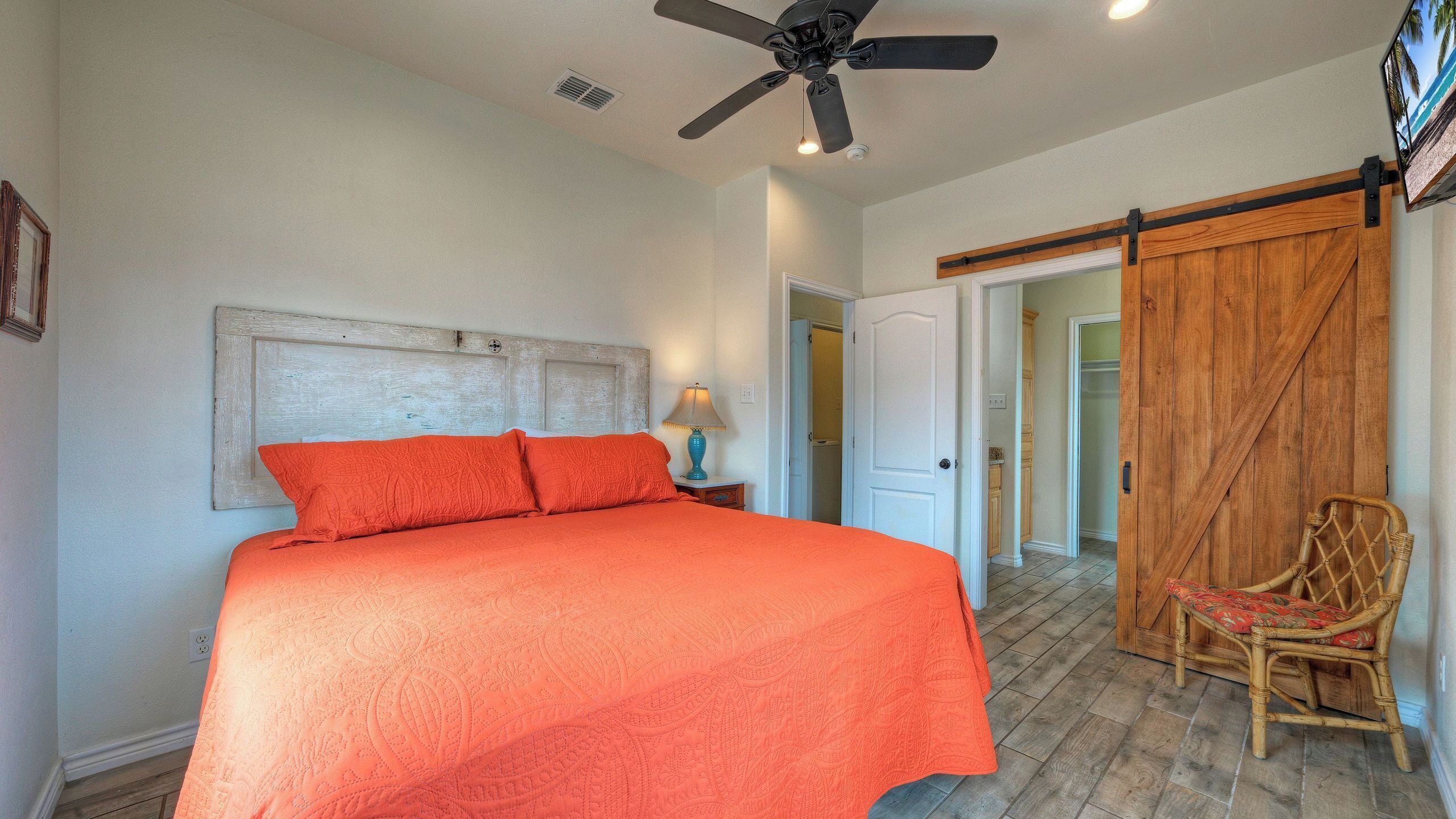 A bedroom with a bed, a ceiling fan, a wooden door, and a wicker chair.