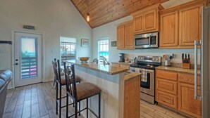 A kitchen with wooden cabinets, a stainless steel refrigerator, a microwave, and a stove. There are bar stools and a dining table. - Cole's Cabin by Avantstay Coastal Retreat Pool Access, Balcony, Water Views + Mins to Beach (Corpus Christi)