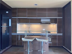 A modern kitchen with dark wood cabinets, a stainless steel refrigerator, a glass-top bar stool, and a built-in oven.