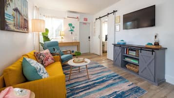 Uma sala de estar com sofá, mesa de centro, TV e área de cozinha com bancada e prateleiras.