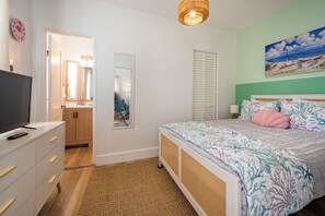 Quarto com cama, televisão em uma cômoda, espelho, porta e um quadro na parede.
