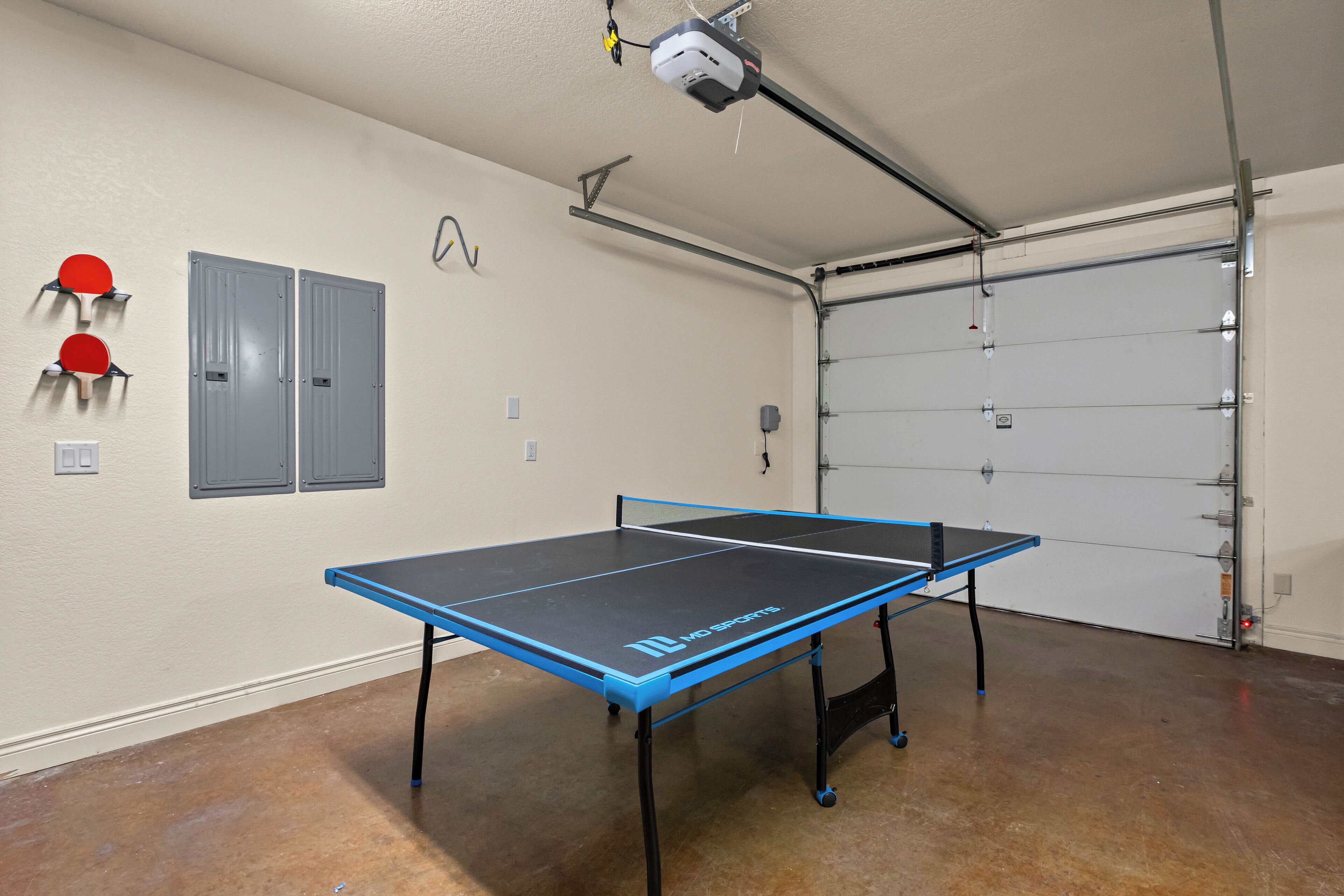 A ping pong table in a garage with a garage door, two grey cabinets, and two red ping pong paddles on the wall.