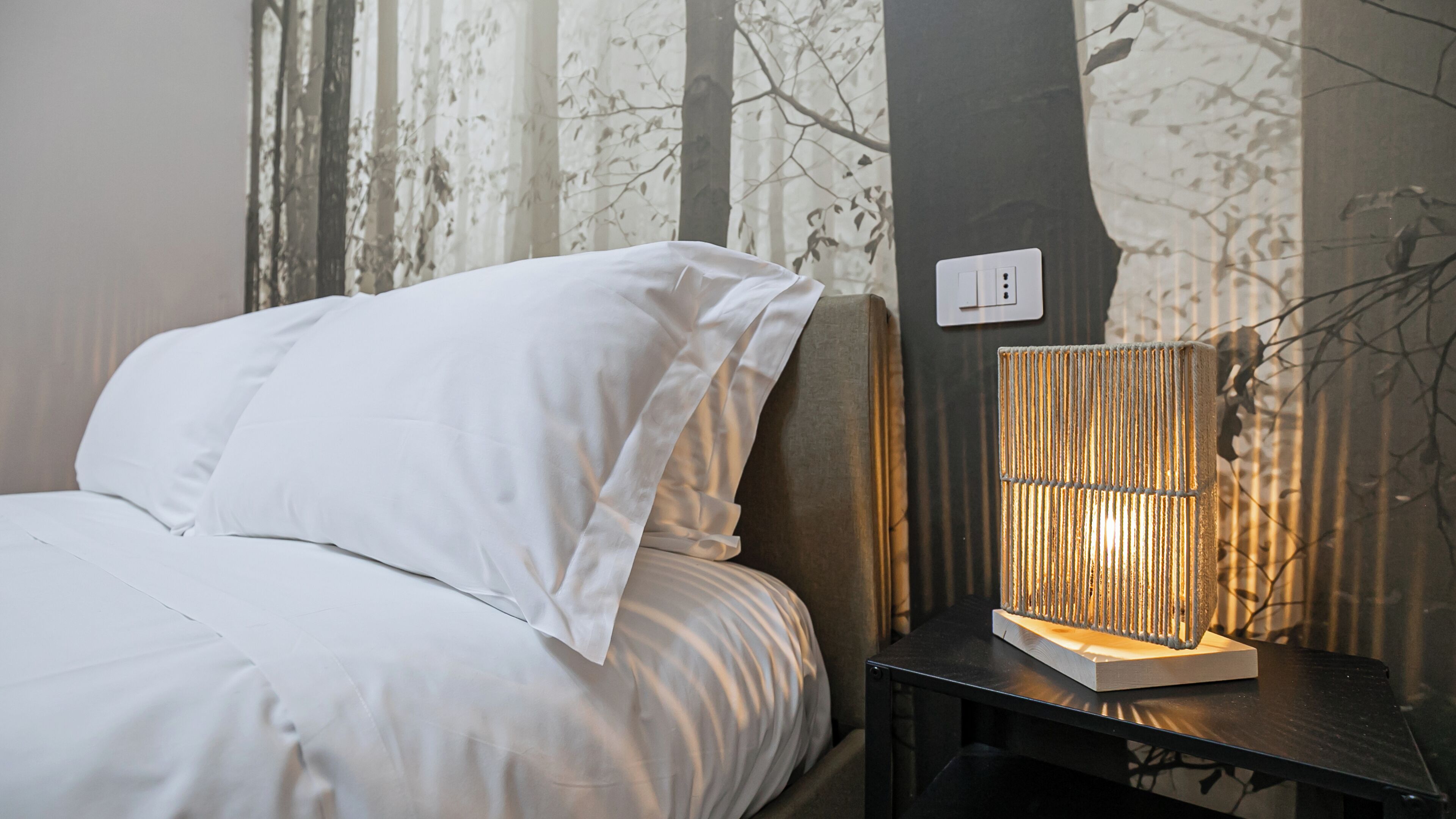 Un lit bien fait, avec du linge de lit blanc, une table de chevet avec une lampe et une prise électrique sur le mur.