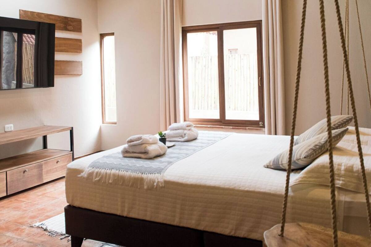 A bedroom with a wooden ceiling, a bed with a hanging swing, a window with curtains, and a small bedside table.