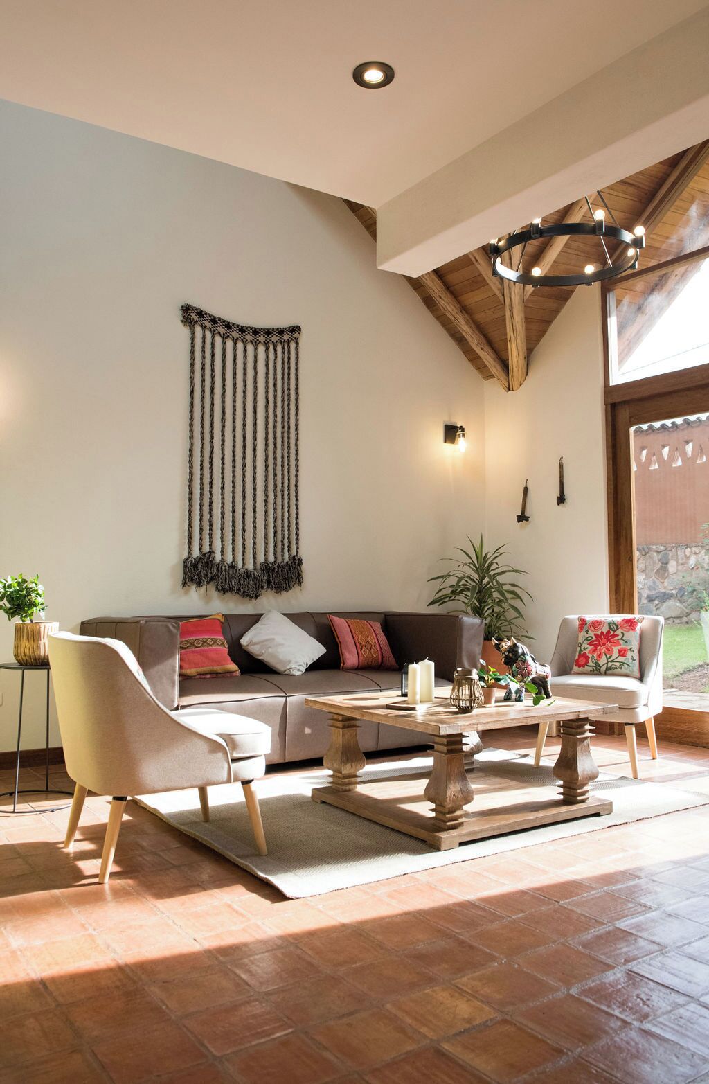 Ampla sala de estar com teto alto, viga de madeira no teto, grande peça de macramé na parede, sofá com almofadas, mesa de centro e uma poltrona.
