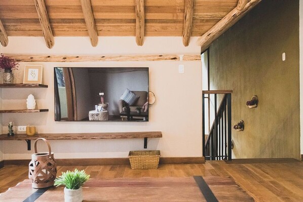 Una sala de estar acogedora con techo de madera, un televisor de pantalla plana grande montado en la pared, un banco de madera, una planta pequeña y una cesta en el suelo.