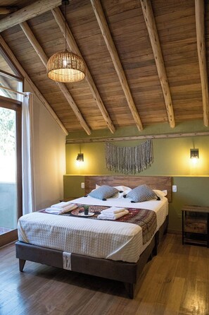 A spacious bedroom with a large bed, wooden ceiling beams, a hanging light fixture, and a wall-mounted lamp. - Killay by Wynwood House (Calca)