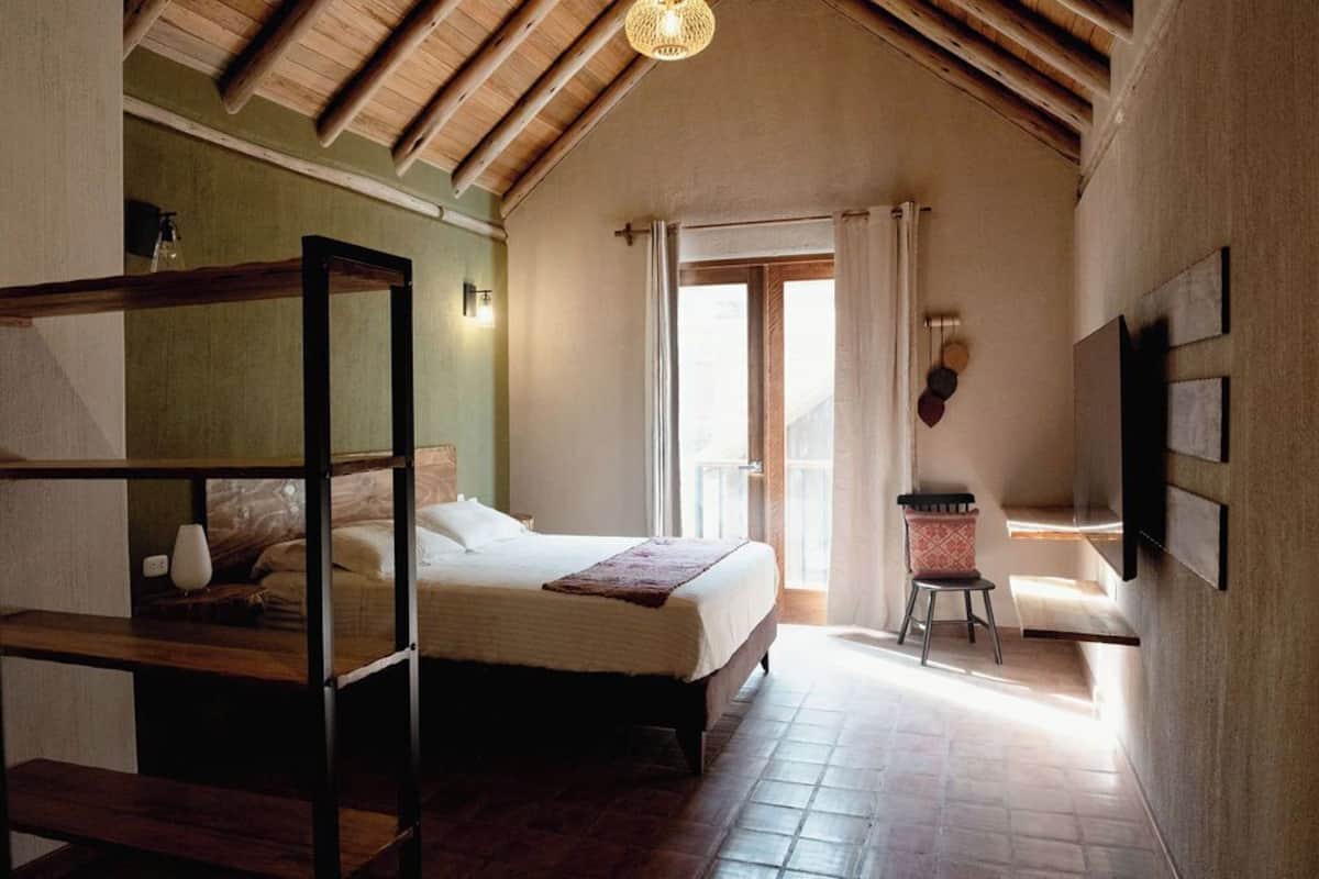 A bedroom with a wooden ceiling, a bed with a white mattress, a small table with a chair, and a window with curtains.