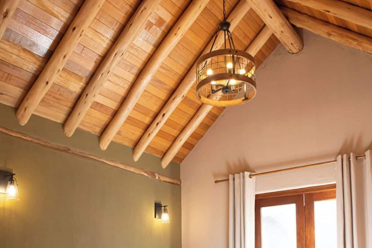 A bedroom with a wooden ceiling, a bed with a headboard, a nightstand, a small table, and a window with curtains.