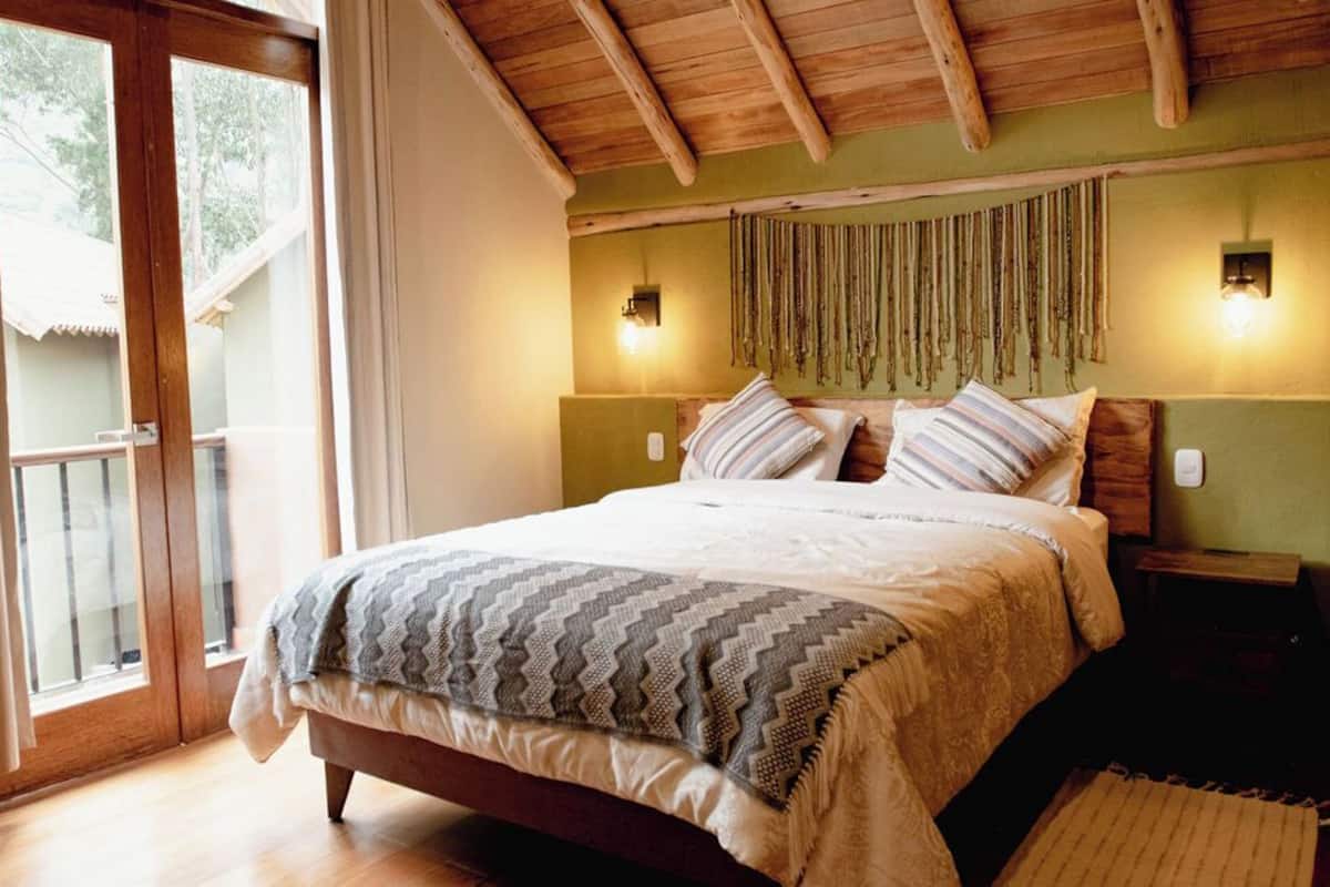 A bedroom with a bed, wooden headboard, two wall-mounted lights, a window with curtains, and a wooden floor.