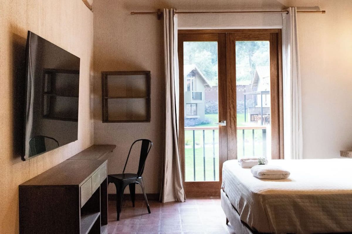 A bedroom with a wooden ceiling, a bed, a desk, a chair, a TV, and a window with a view of buildings.