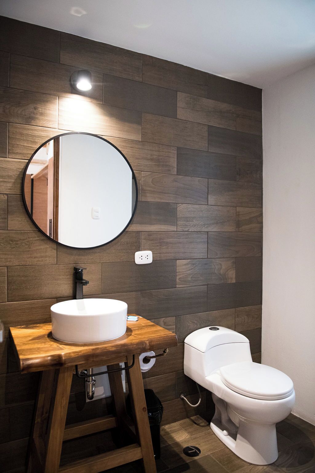 A modern bathroom with a round mirror, wooden vanity, white sink, and toilet.