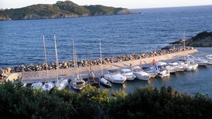 En småbåtshamn med båtar förtöjda längs en klippig strand, omgiven av grönska och en avlägsen ö.