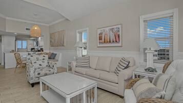 A spacious living area with a sofa, coffee table, and wicker chairs. A kitchen is visible in the background.