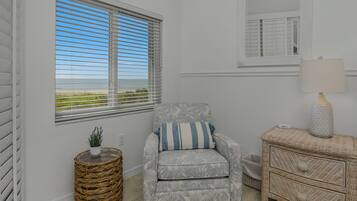 Ein Zimmer mit Meerblick durch ein Fenster mit Jalousien, ein Korbstuhl mit einer Pflanze, ein gemustertes Sessel mit einem Kissen und eine Korbmöbelkommode mit einer Lampe.