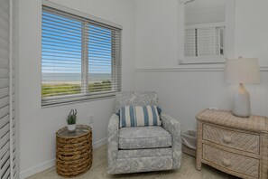 Una habitación con vistas al mar a través de una ventana con persianas, una mesita de mimbre con una planta, un sillón estampado con un cojín y un aparador de mimbre con una lámpara.