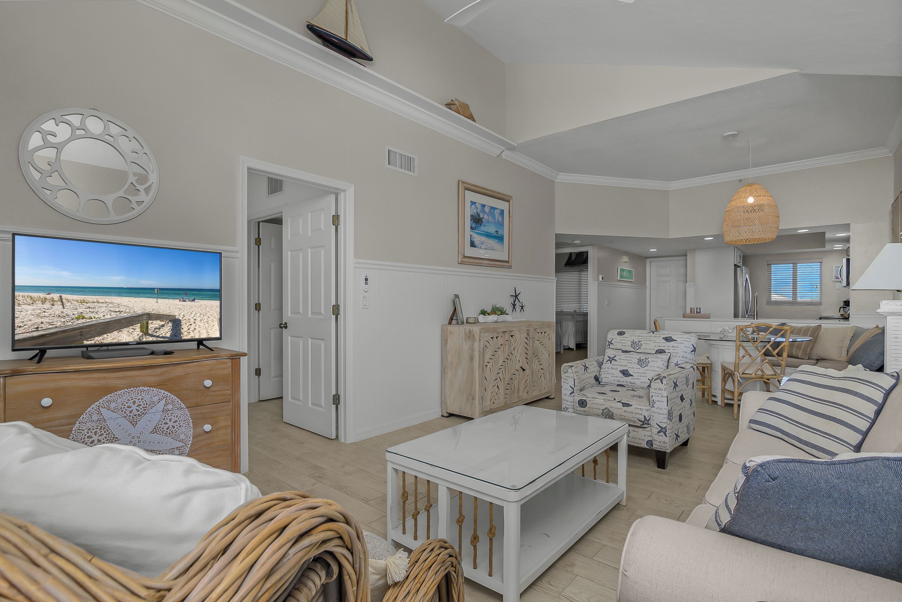 Ein Wohnzimmer mit Fernseher, der eine Strandansicht zeigt, eine Essecke mit Tisch und Stühlen sowie eine Küche im Hintergrund.