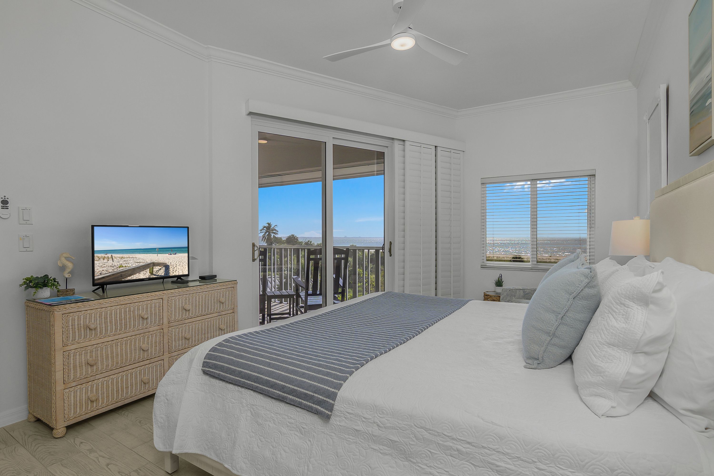 A bedroom with a bed, a television, a balcony with a view of the beach, and a ceiling fan.