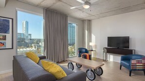 Een moderne woonkamer met een grijze bank, gele kussens, een houten salontafel met wielen, een blauwe fauteuil, een televisie op een meubel en een stadszicht door grote ramen.