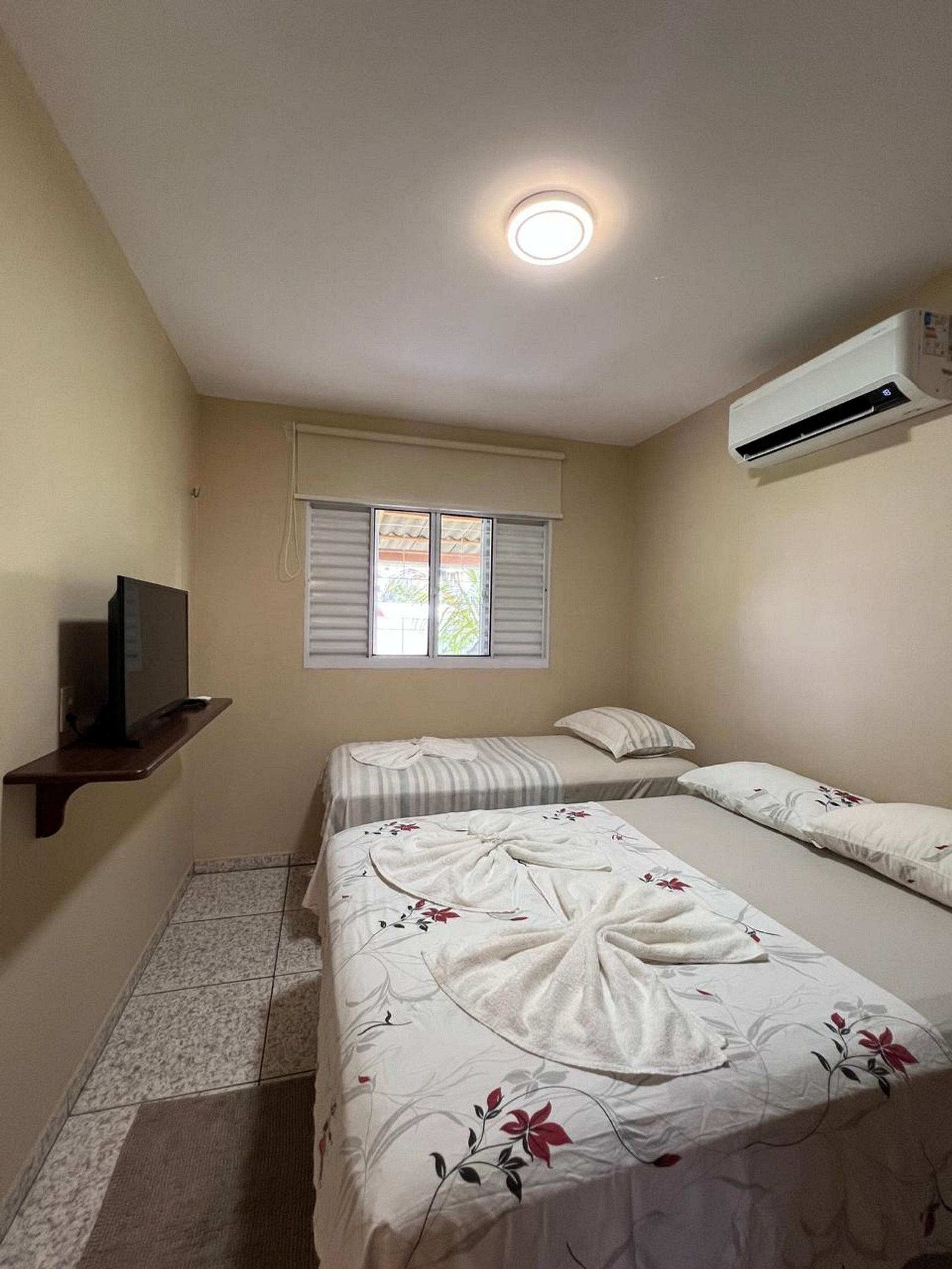 A bedroom with two beds, a TV on a shelf, a window with blinds, and an air conditioning unit.