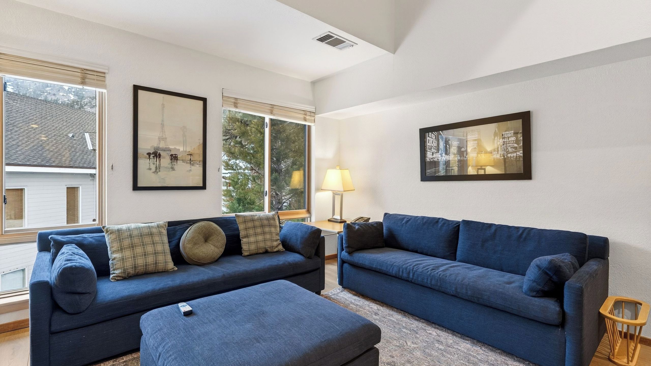 A living room with two blue sofas, a coffee table, and a lamp. There are framed pictures on the walls and a window with blinds.